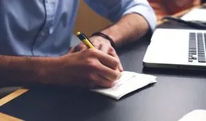Man writing notes beside laptop