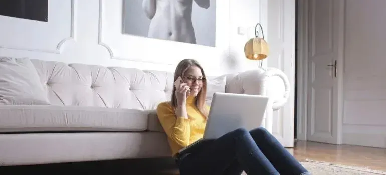 Woman in yellow sweater on phone using laptop by sofa