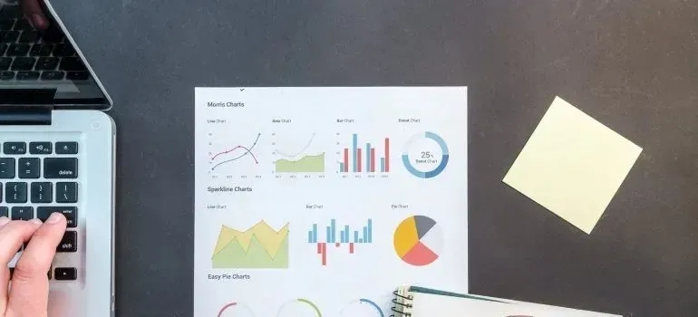 Hand at laptop beside printed charts and sticky note