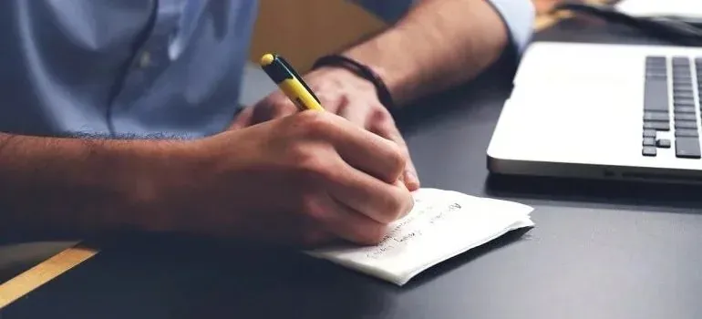 Hands writing notes beside laptop on dark desk