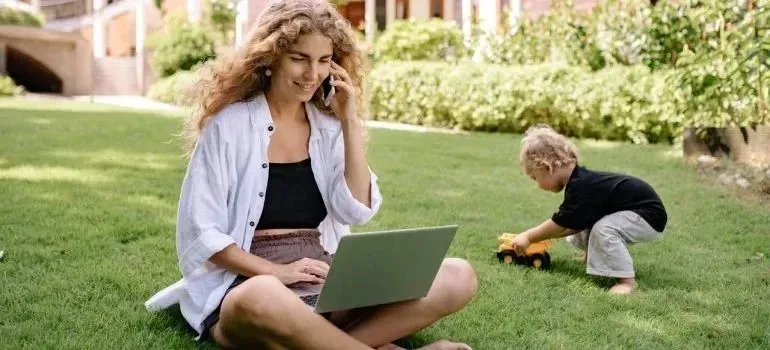 Woman with laptop and phone on lawn, child playing nearby
