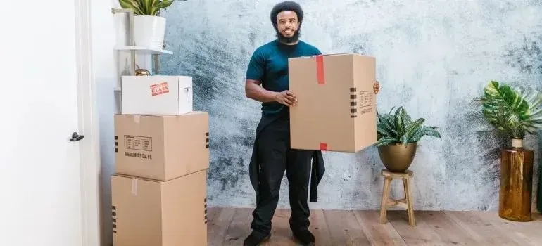 Smiling mover holding large box beside stacked boxes and plants