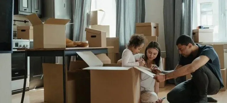 Family labeling open moving box in apartment full of boxes