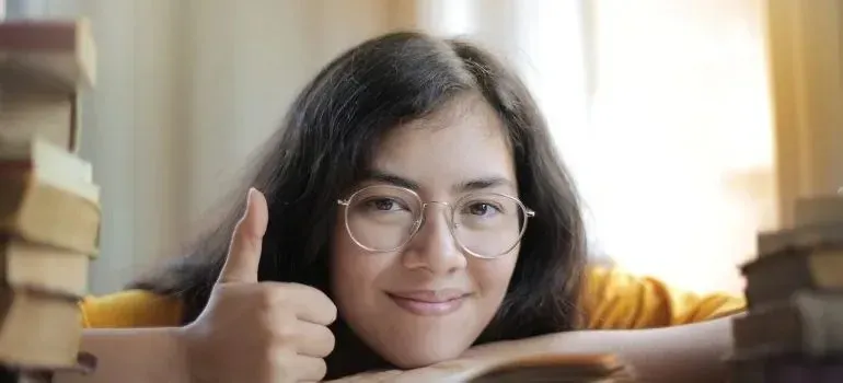Smiling girl in glasses giving thumbs up near books