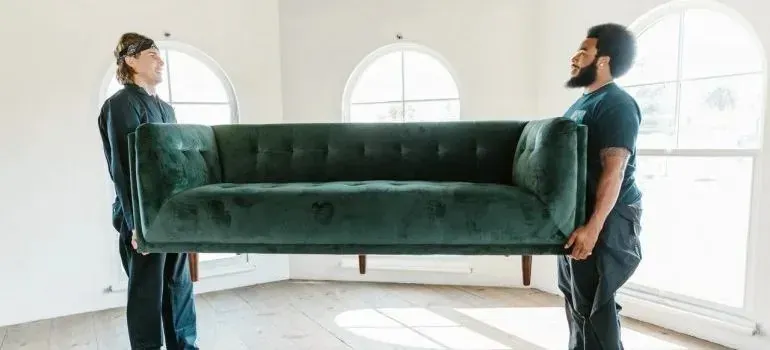 Two movers carrying green velvet sofa in bright room