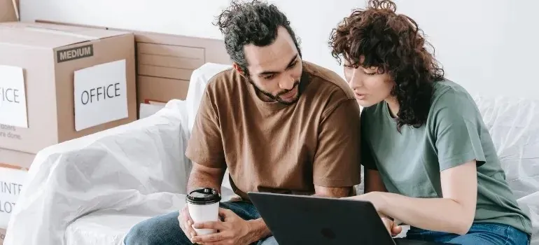 Couple with laptop on couch among office-labeled moving boxes
