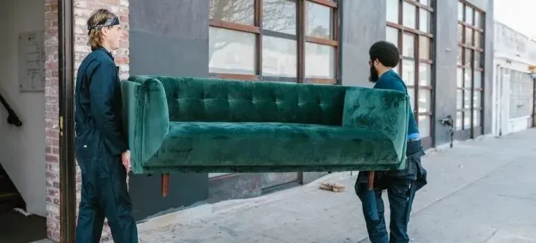 Two movers in plain workwear carrying green velvet sofa