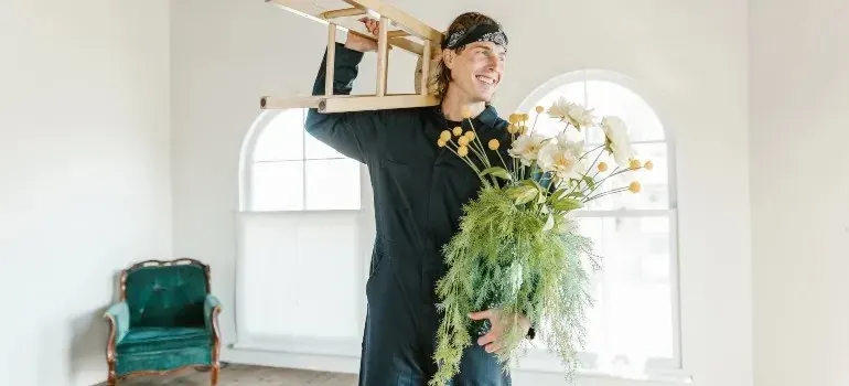 Cheerful mover carrying wooden stool and flowers indoors
