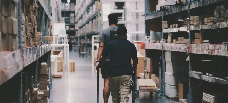 Two men pushing cart through warehouse shelving aisles