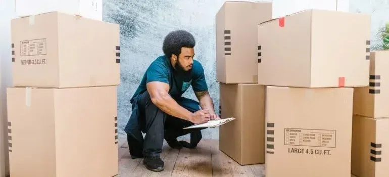Mover with clipboard crouching among labeled cardboard moving boxes