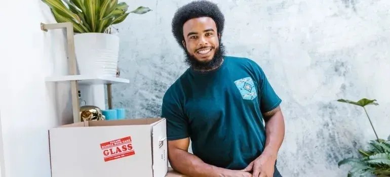 Smiling mover beside glass-labeled moving box and houseplants