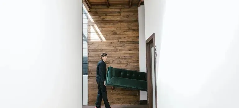 Person carrying green velvet sofa through wood-paneled hallway