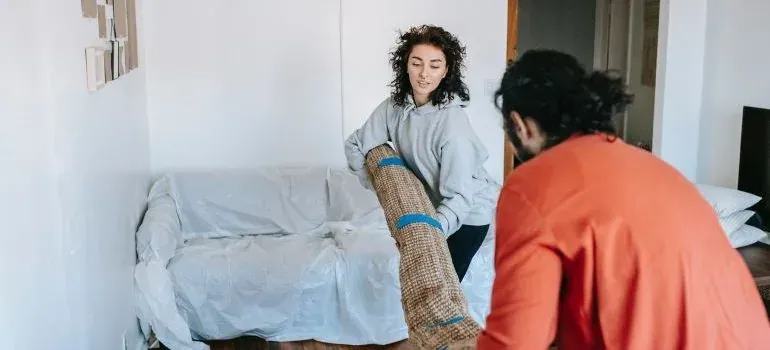Couple carrying rolled rug beside plastic-covered sofa