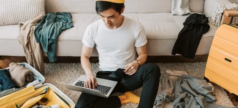 Man with laptop packing suitcase and clothes on floor