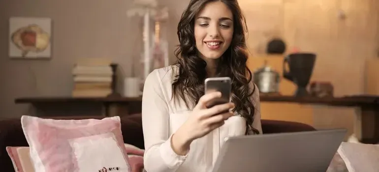 Smiling woman with phone and laptop on cozy sofa