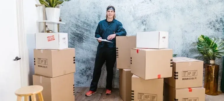 Mover with clipboard standing among stacked moving boxes indoors