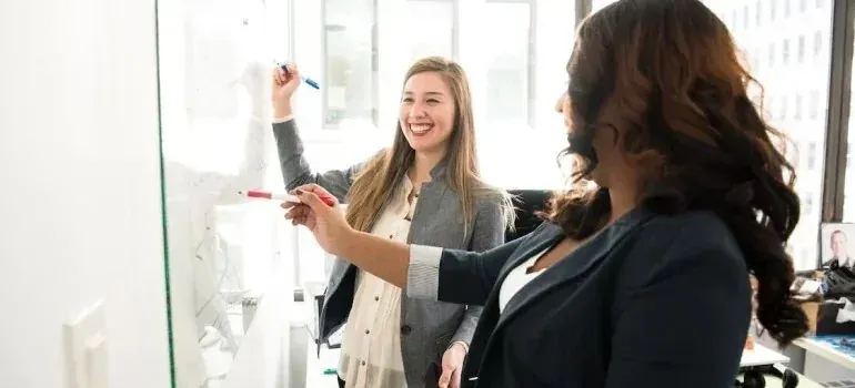 Two smiling businesswomen writing on office whiteboard