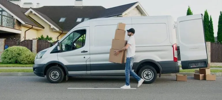 Man carrying stacked boxes to white van on suburban street
