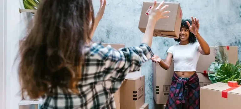 Two women playfully tossing cardboard box while packing