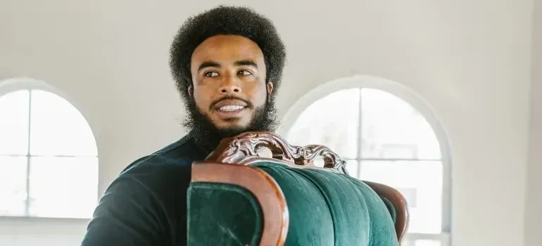 Smiling man carrying green vintage armchair in bright room