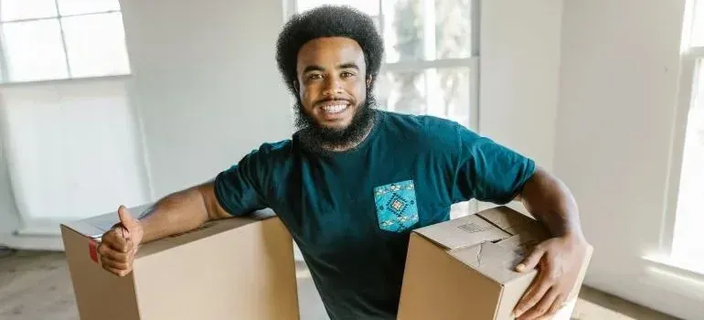 Smiling bearded mover holding two cardboard boxes indoors