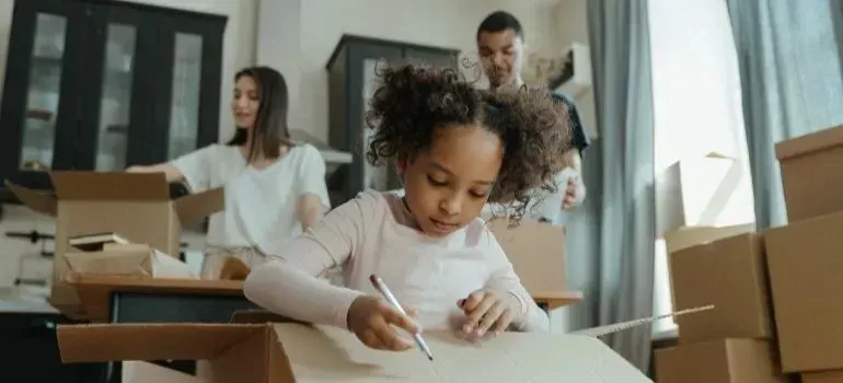 Girl drawing on cardboard box while family unpacks