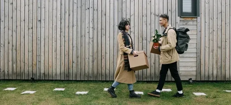 Couple carrying box and plant past wooden house facade