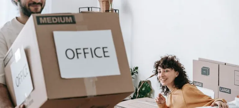Man carrying box labeled OFFICE while woman watches