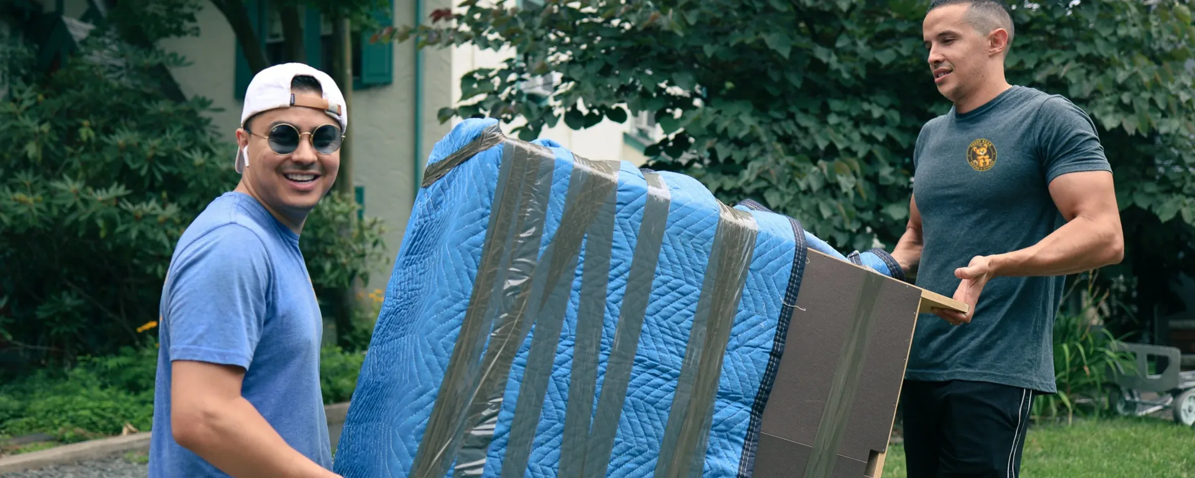 Two Teddy Bear Moving crew members carrying a wrapped piece of furniture across a front lawn